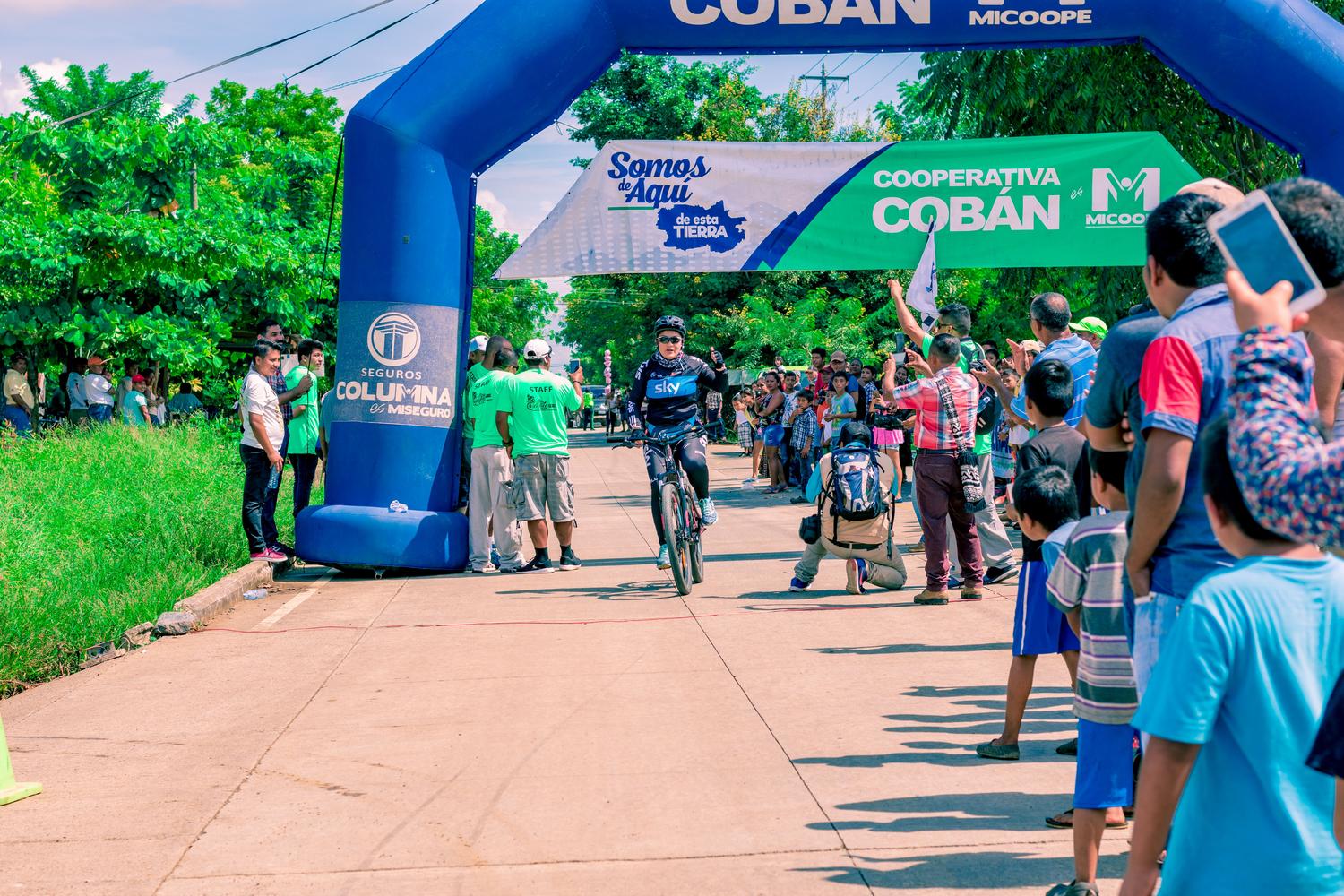 Ciclista cruzando la línea de meta en una carrera al aire libre con el público animando.