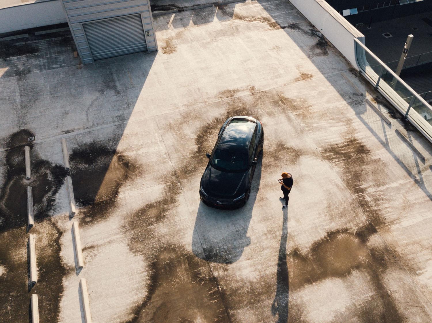 Vista aérea de un coche aparcado en un parking exterior con una persona al lado consultando su móvil.