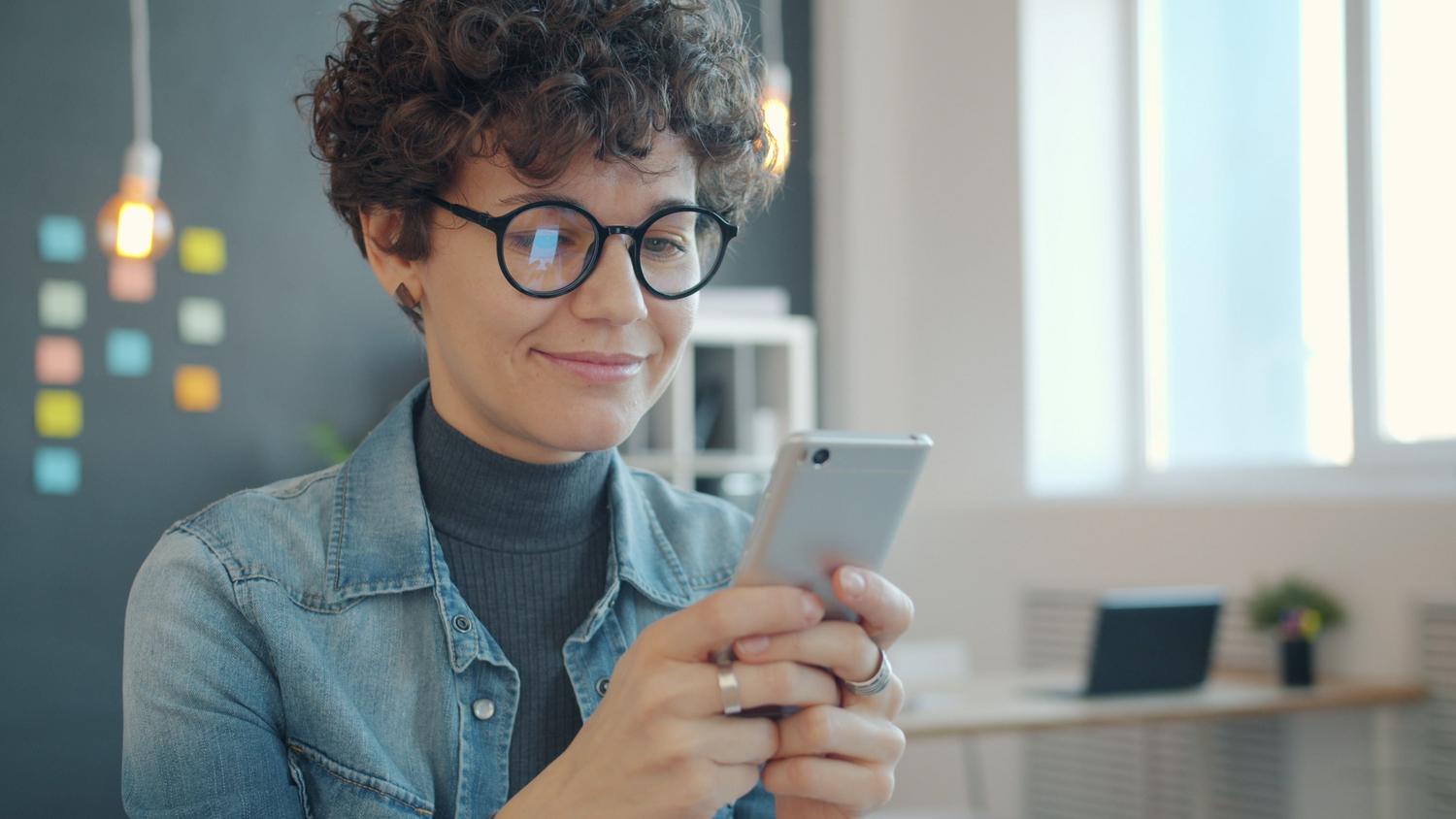 Mujer joven con gafas utilizando un smartphone en una oficina moderna y luminosa.