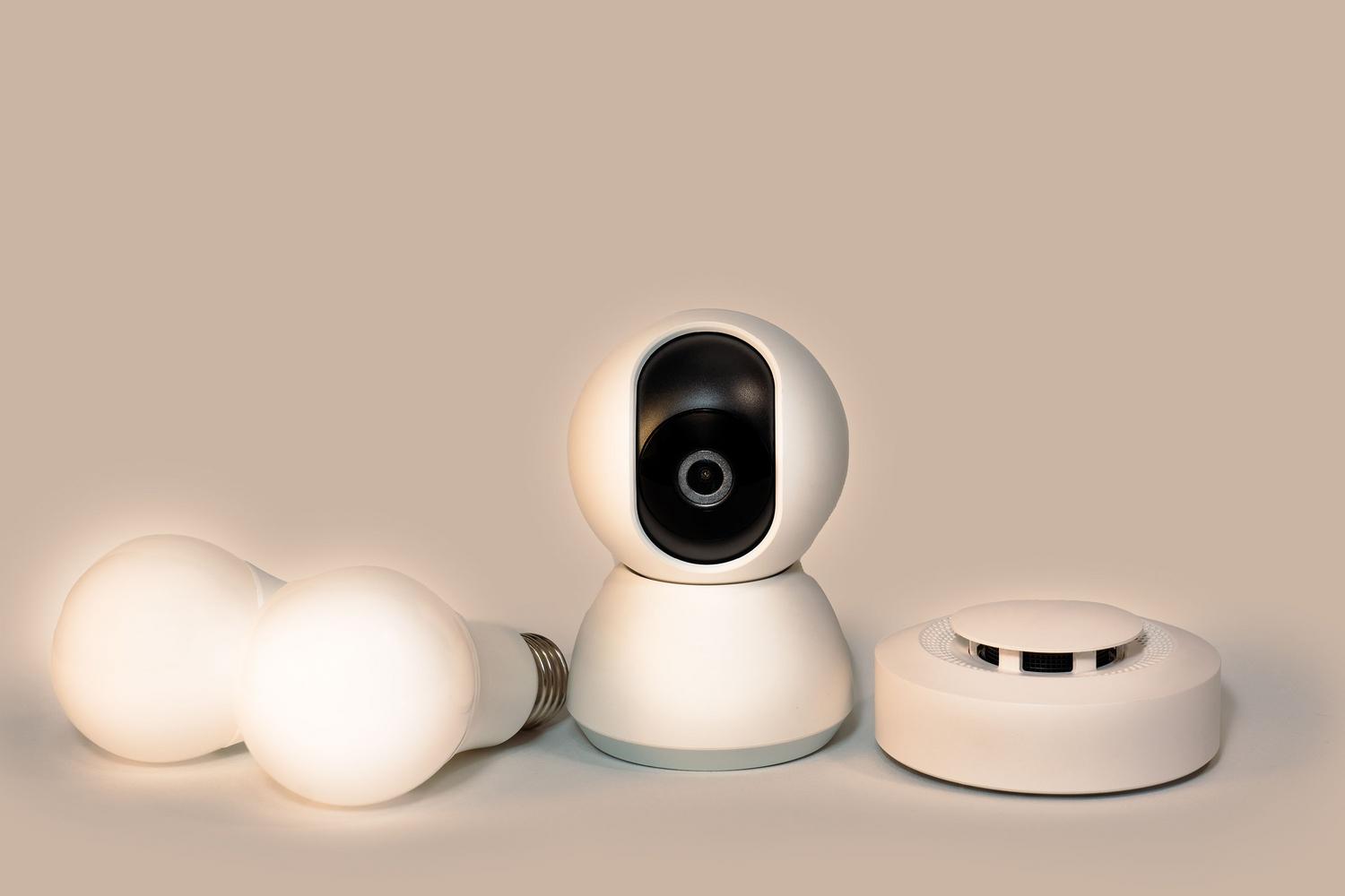 Conjunto de dispositivos inteligentes incluyendo bombillas LED, una cámara de vigilancia y un detector de humo sobre un fondo neutro.